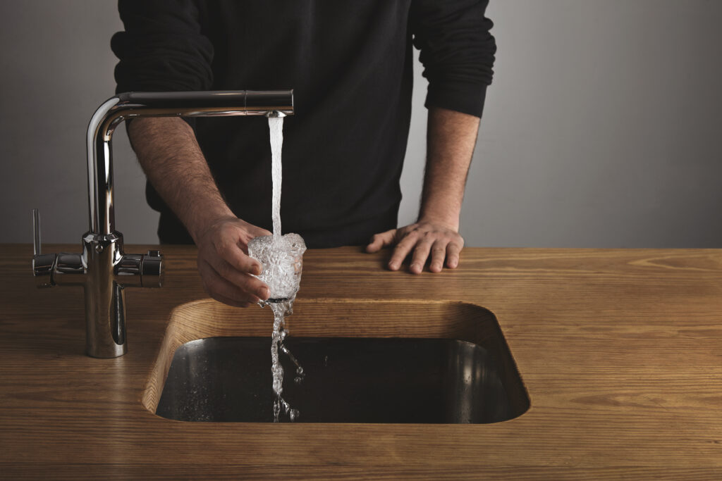 An image of a persons body with their sleeves rolled up filling a small glass of water up over a silver kitchen faucet